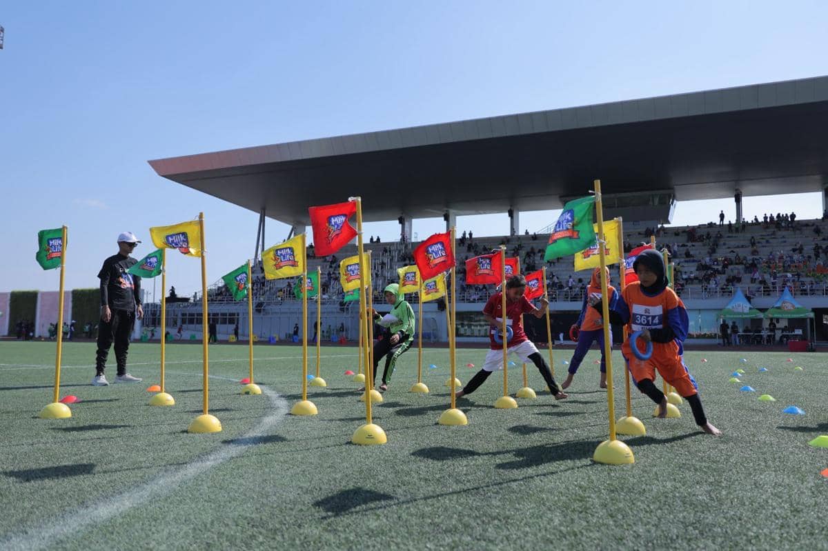 Anak-anak berpartisipasi dalam lomba lari rintangan dengan bendera warna-warni di ajang MilkLife Athletics Challenge Seri 1 2026 di stadion.