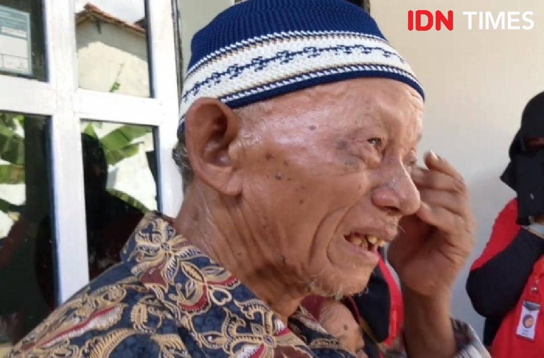 Seorang pria lanjut usia mengenakan peci rajut biru dan batik menangis haru di depan rumah setelah menerima kunci rumah baru.