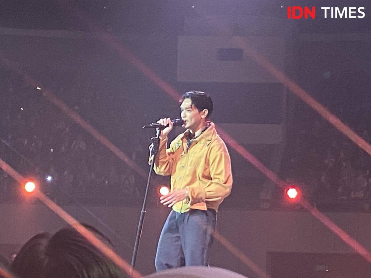 Seorang penyanyi mengenakan jaket kuning sedang bernyanyi di atas panggung konser dengan pencahayaan merah di Plenary Hall JICC Jakarta.