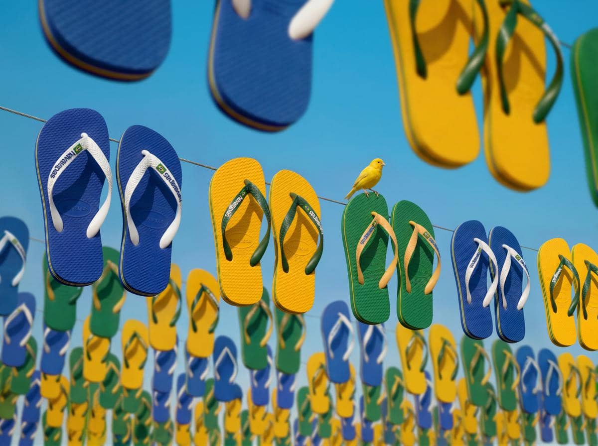 Deretan sandal Havaianas berwarna biru, hijau, dan kuning tergantung di tali dengan latar langit biru dan seekor burung kuning di atasnya.