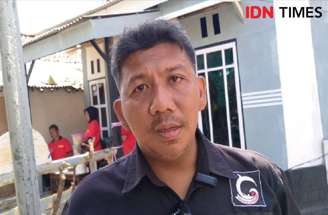 Seorang pria mengenakan kemeja hitam berlogo komunitas berdiri di depan rumah dengan beberapa orang di latar belakang.