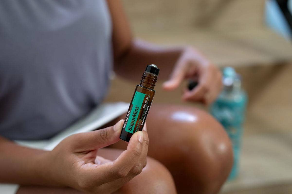 Seseorang memegang botol minyak angin roll-on berlabel SuperMint di tangan, dengan latar belakang kaki dan botol minum di dekatnya.