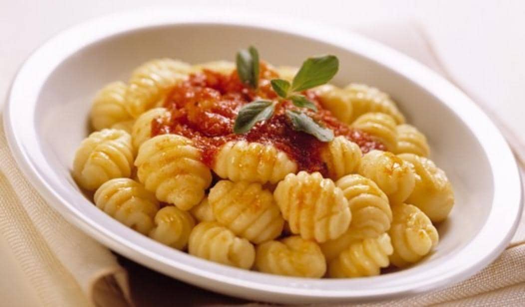 Semangkuk ñoquis lembut disajikan dengan saus tomat merah di atasnya dan hiasan daun basil segar di piring putih.