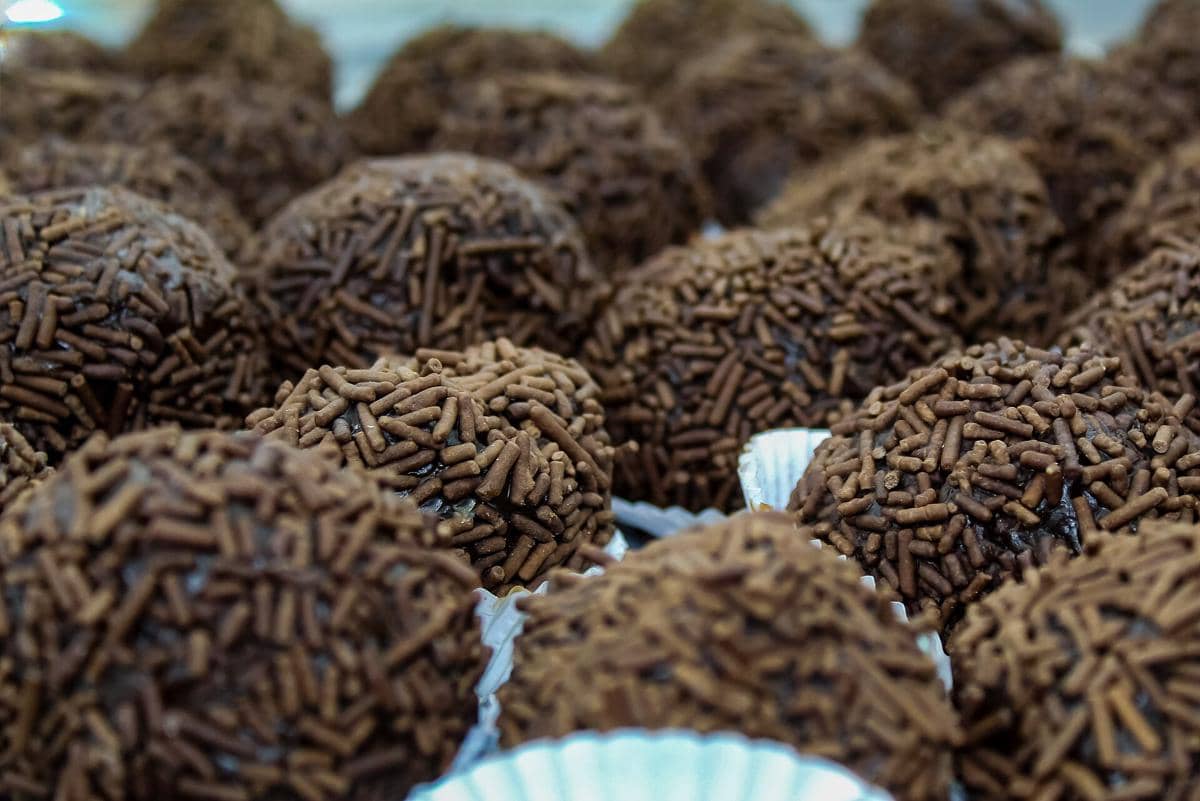 Ilustrasi brigadeiro berbentuk bulat dengan lapisan meses cokelat, disajikan dalam wadah kertas kecil.
