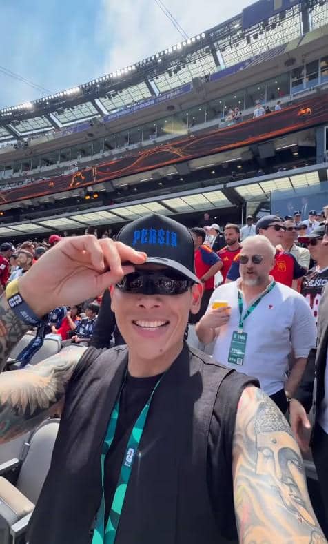 Seorang pria bertopi hitam dan berkacamata hitam tersenyum sambil berfoto selfie di stadion yang dipenuhi penonton pertandingan sepak bola.