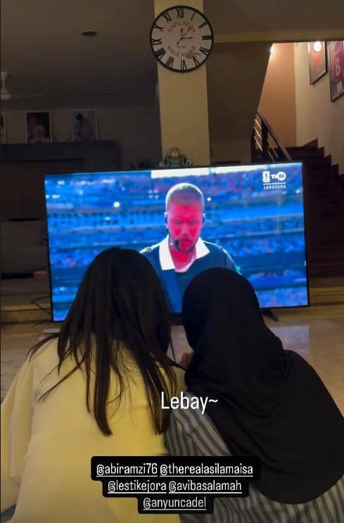 Dua orang duduk di lantai ruang tamu menonton penampilan di televisi besar yang menayangkan acara Piala Dunia.