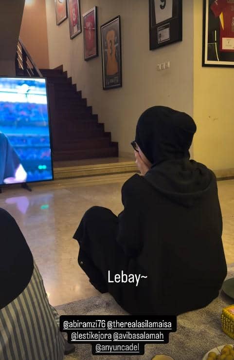 Seorang perempuan berhijab duduk di lantai ruang tamu menonton televisi yang menayangkan acara musik, dengan tangga dan bingkai foto di belakangnya.