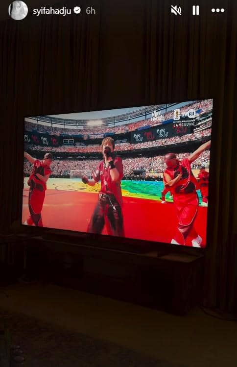 Layar televisi menampilkan penampilan panggung berwarna merah di stadion besar dengan penyanyi dan penari mengenakan kostum merah.