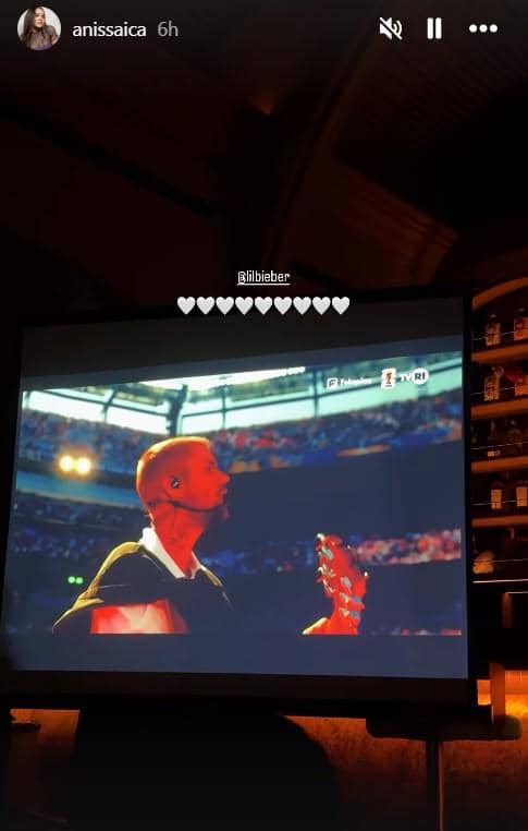 Layar proyektor menampilkan penampilan musisi di stadion besar dengan pencahayaan panggung dan penonton memenuhi tribun.