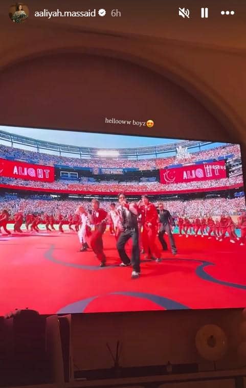 Layar televisi menampilkan penampilan panggung dengan sekelompok penari berpakaian merah di stadion besar yang dipenuhi penonton.