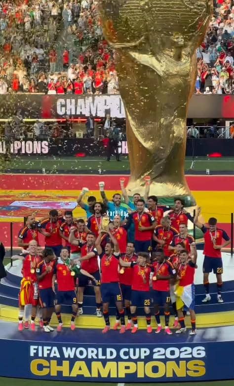 Tim sepak bola mengenakan seragam merah dan biru merayakan kemenangan di podium dengan trofi besar Piala Dunia 2026 di stadion penuh penonton.