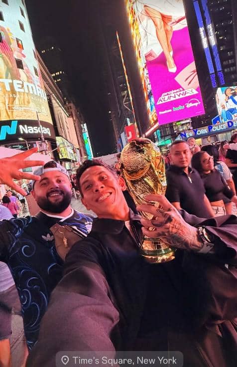 Seorang pria berpose sambil memegang trofi Piala Dunia di tengah keramaian malam di Times Square, New York, dengan latar papan reklame terang.