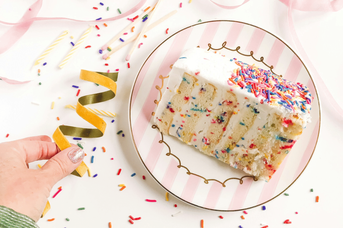 Potongan kue ulang tahun rasa vanila dengan taburan sprinkle warna-warni di atas piring bergaris merah muda dan putih.