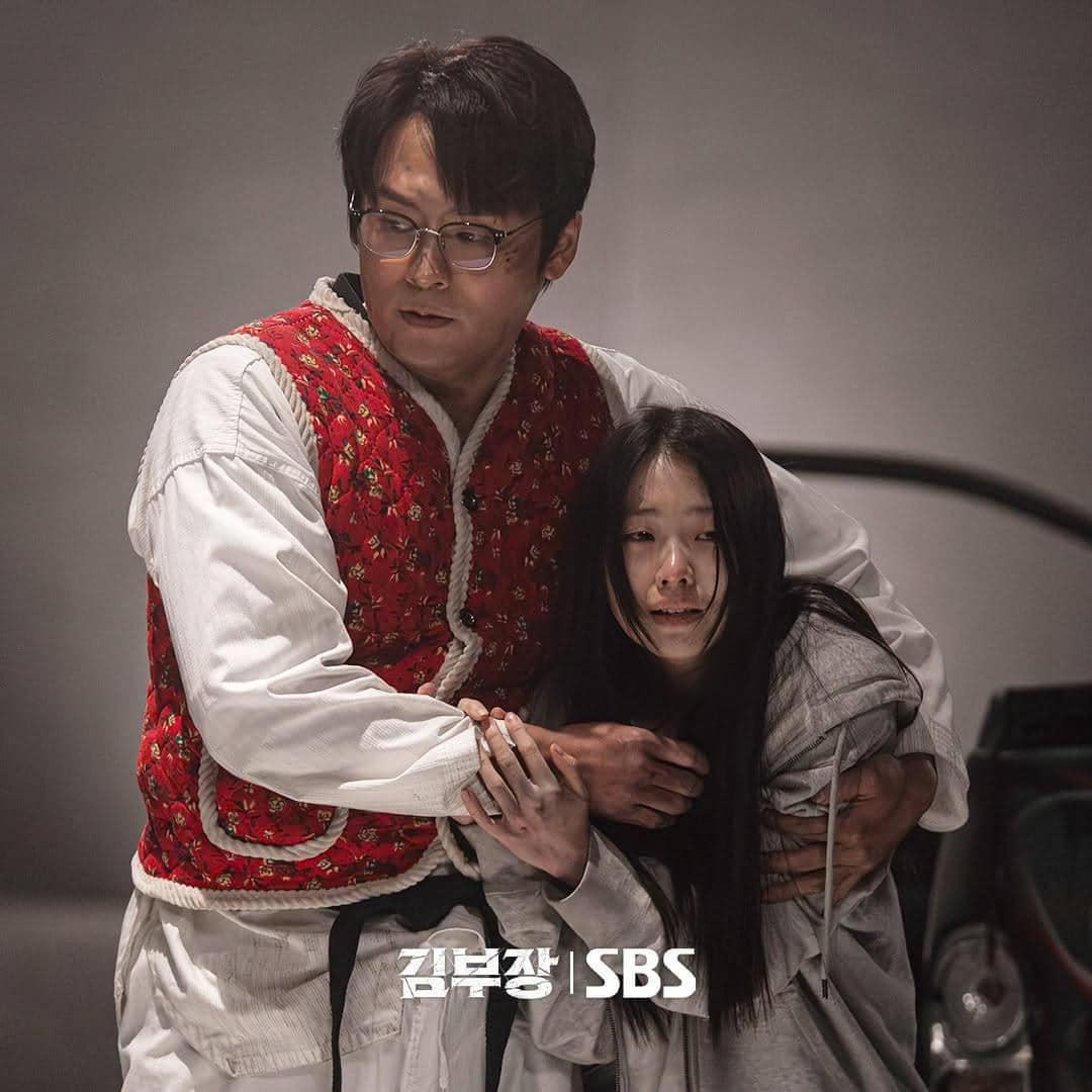 Seorang pria mengenakan rompi merah bermotif bunga memegang seorang wanita berambut panjang yang tampak ketakutan di adegan drama Korea Agent Kim Reactivated.