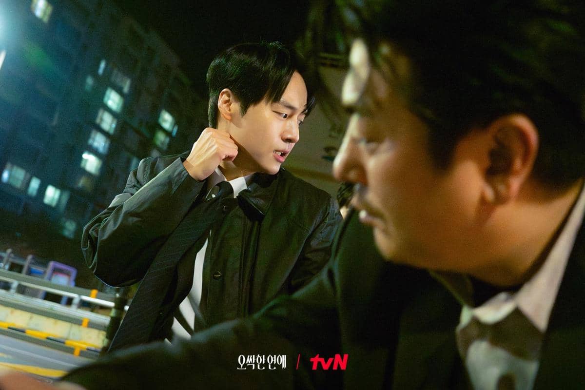 Dua pria mengenakan jas hitam sedang berbicara serius di malam hari dengan latar gedung apartemen dalam adegan drama Spooky in Love.