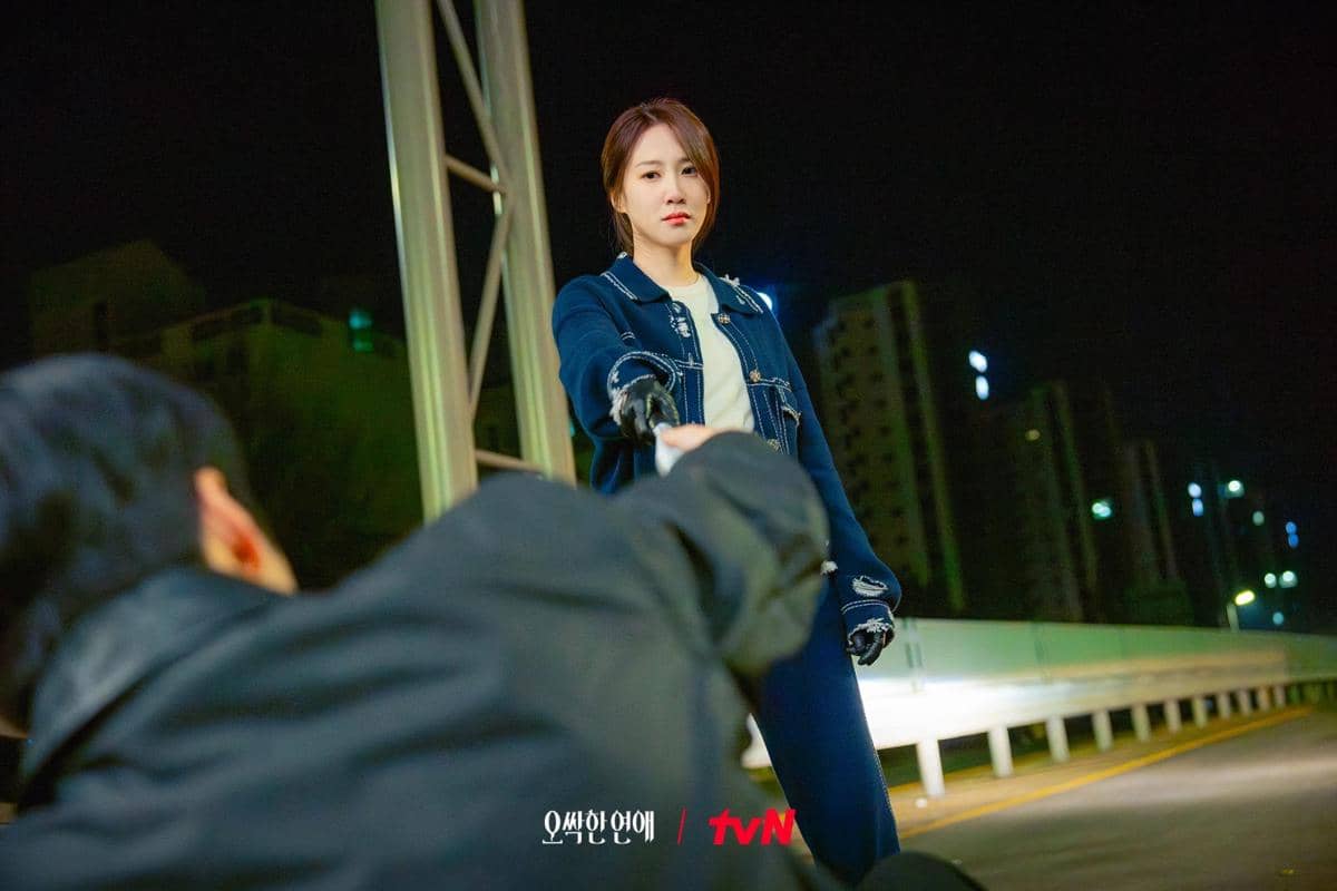 Seorang wanita berdiri di jalan layang pada malam hari mengenakan jaket biru, menatap seseorang yang terjatuh di depannya.