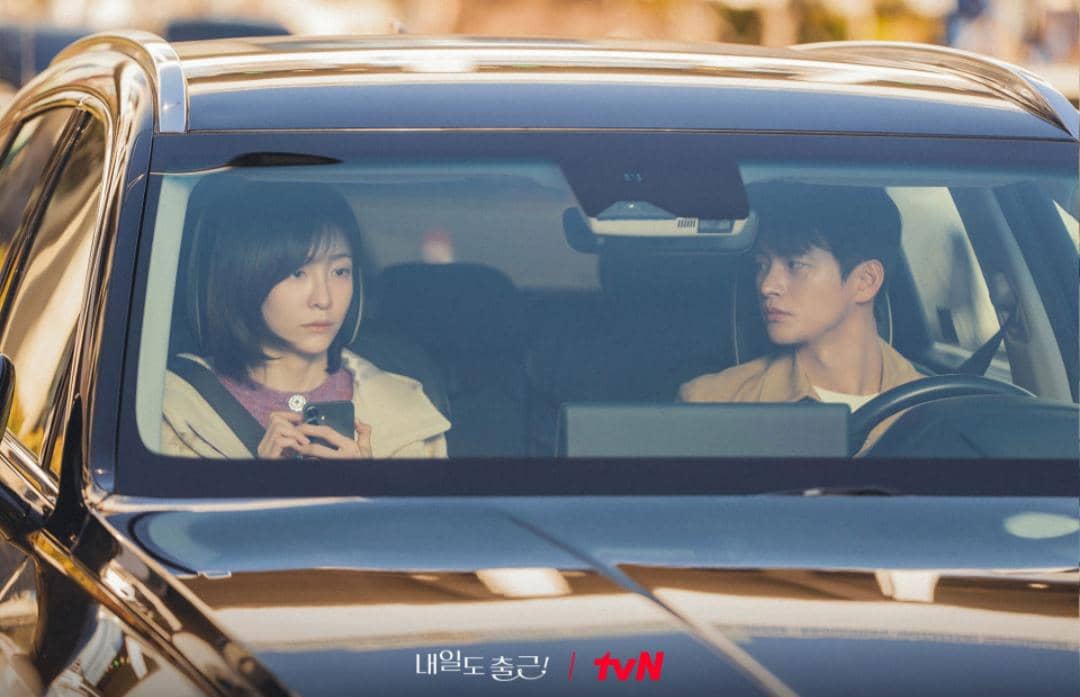 Dua karakter duduk di dalam mobil, satu memegang ponsel sementara yang lain menatapnya serius dalam adegan drama See You at Work Tomorrow.