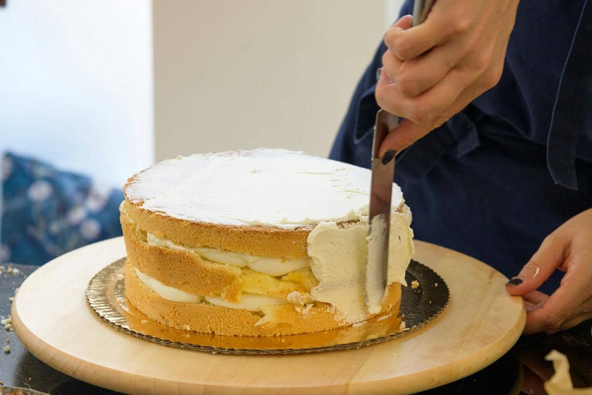 Seseorang sedang mengoleskan krim putih pada lapisan kue bolu di atas alas kayu bundar menggunakan spatula logam.
