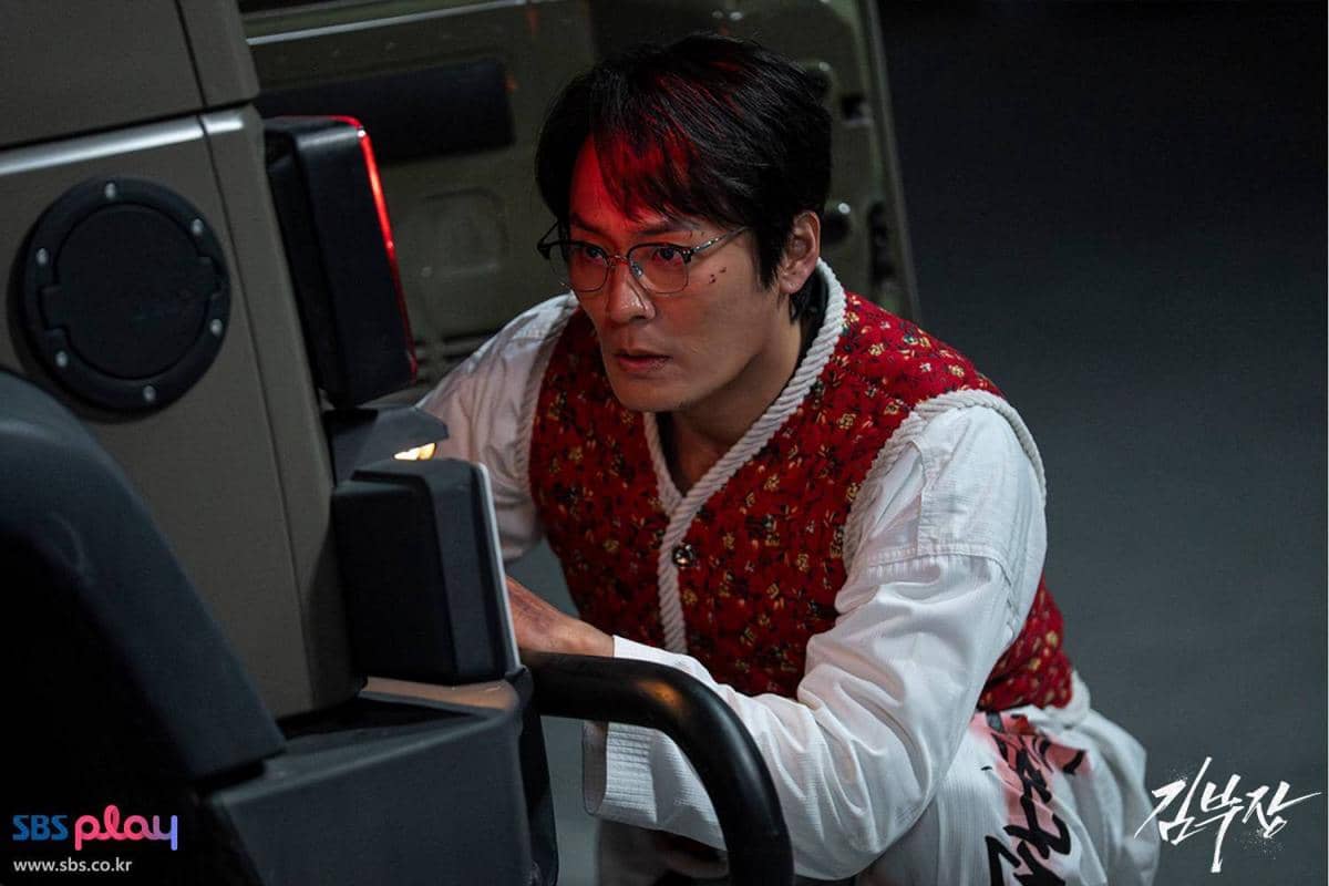 Seorang pria berkacamata mengenakan rompi merah bermotif sedang berjongkok di samping kendaraan dalam adegan drama Korea Agent Kim Reactivated.