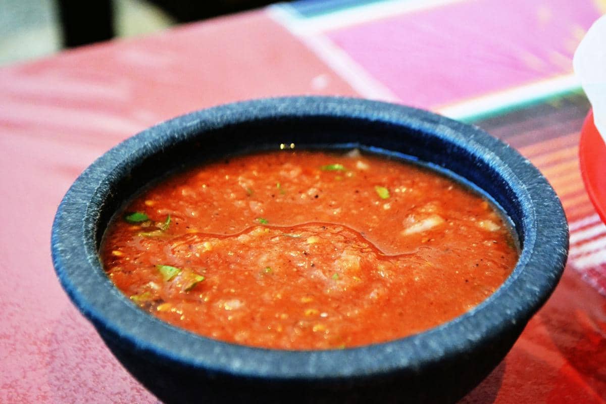 Semangkuk salsa merah khas restoran disajikan dalam mangkuk batu hitam di atas meja dengan taplak berwarna cerah.