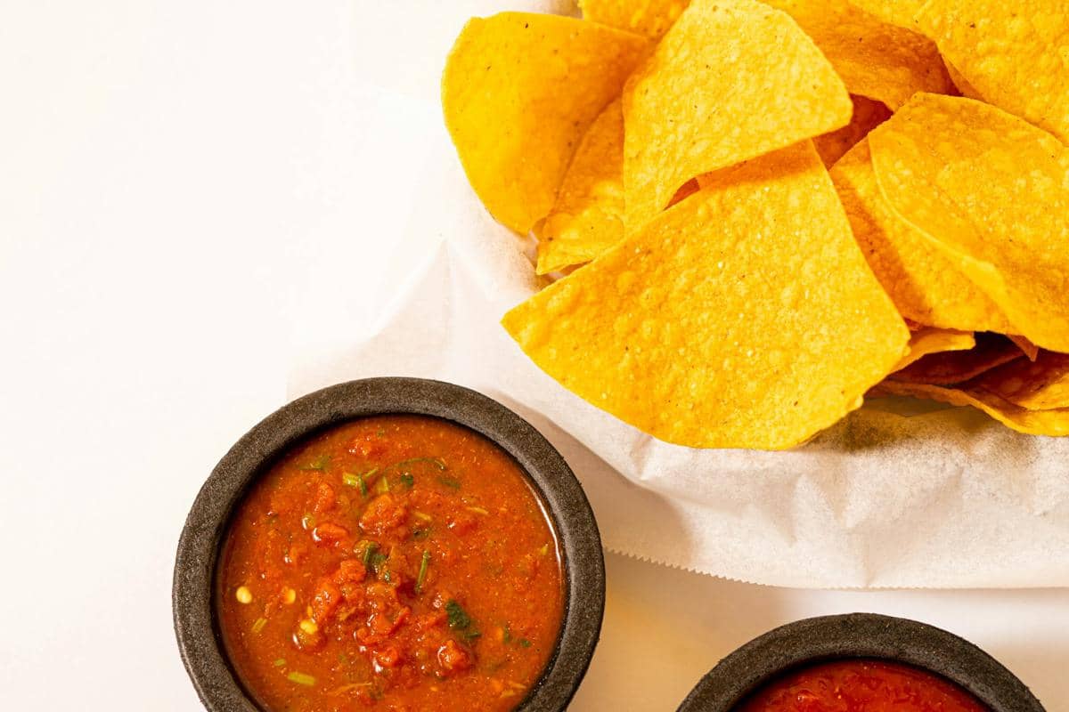 Semangkuk saus salsa merah dengan potongan cabai dan rempah disajikan bersama keripik tortilla jagung renyah di atas meja putih.