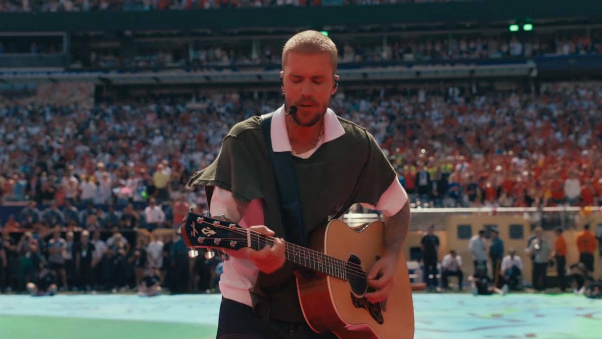 Seorang penyanyi tampil di atas panggung stadion besar sambil memainkan gitar akustik di depan penonton yang memenuhi tribun.
