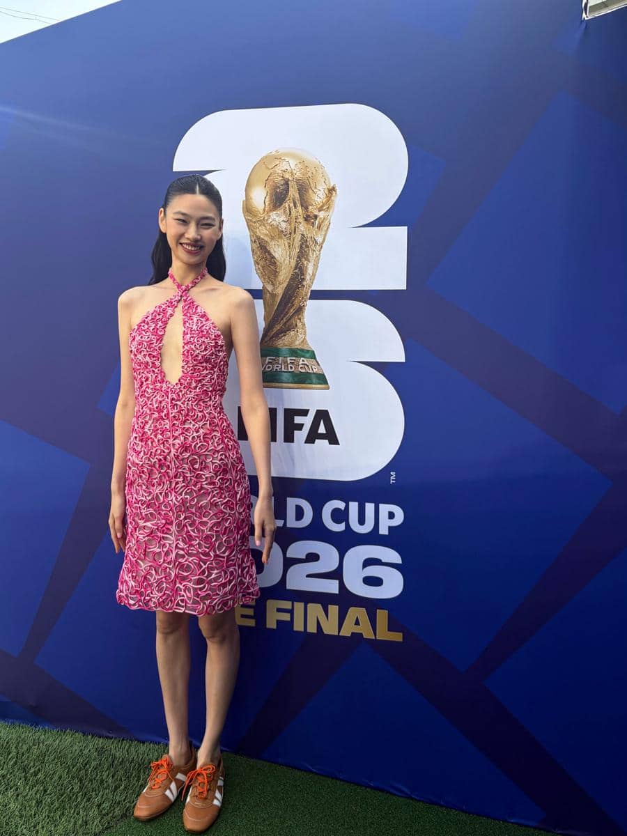 Seorang perempuan mengenakan gaun merah muda bermotif berdiri di depan latar bertuliskan FIFA World Cup 2026 The Final dengan trofi Piala Dunia.