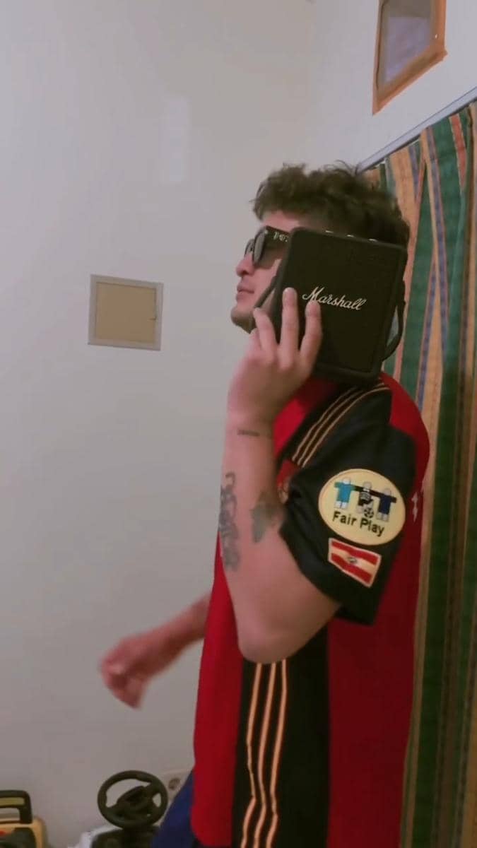 Seorang pria mengenakan jersey tim nasional Spanyol memegang speaker Marshall di telinganya sambil mengenakan kacamata hitam di dalam ruangan.