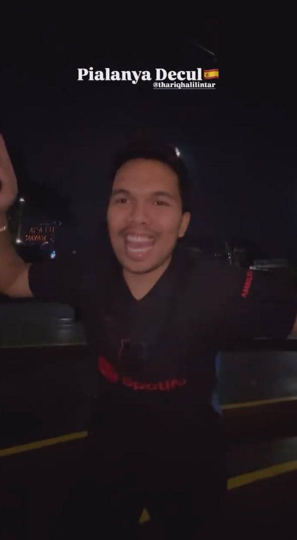 Seorang pria mengenakan kaus hitam tersenyum lebar dan mengangkat tangan saat malam hari dengan tulisan 'Pialanya Decul' di atasnya.