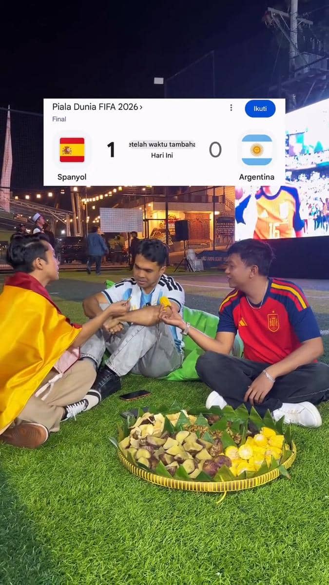 Tiga pria mengenakan jersey Spanyol dan Argentina menonton final Piala Dunia 2026 sambil duduk di rumput dengan hidangan tradisional di depan mereka.