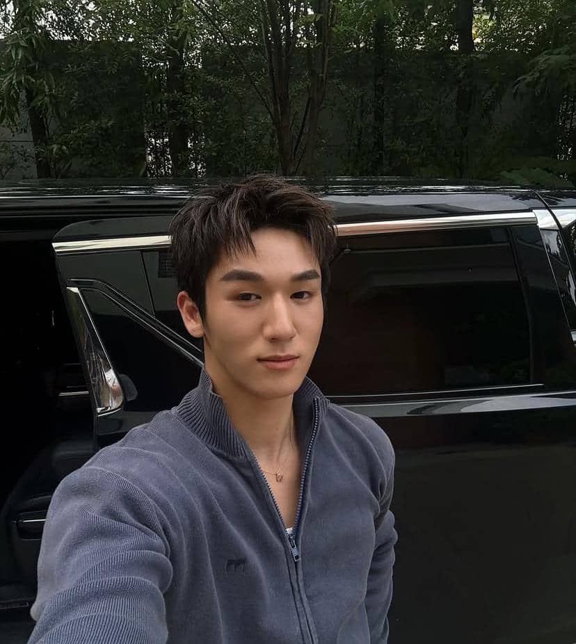 Seorang pria mengenakan jaket abu-abu berfoto selfie di depan mobil hitam dengan latar pepohonan hijau di belakangnya.