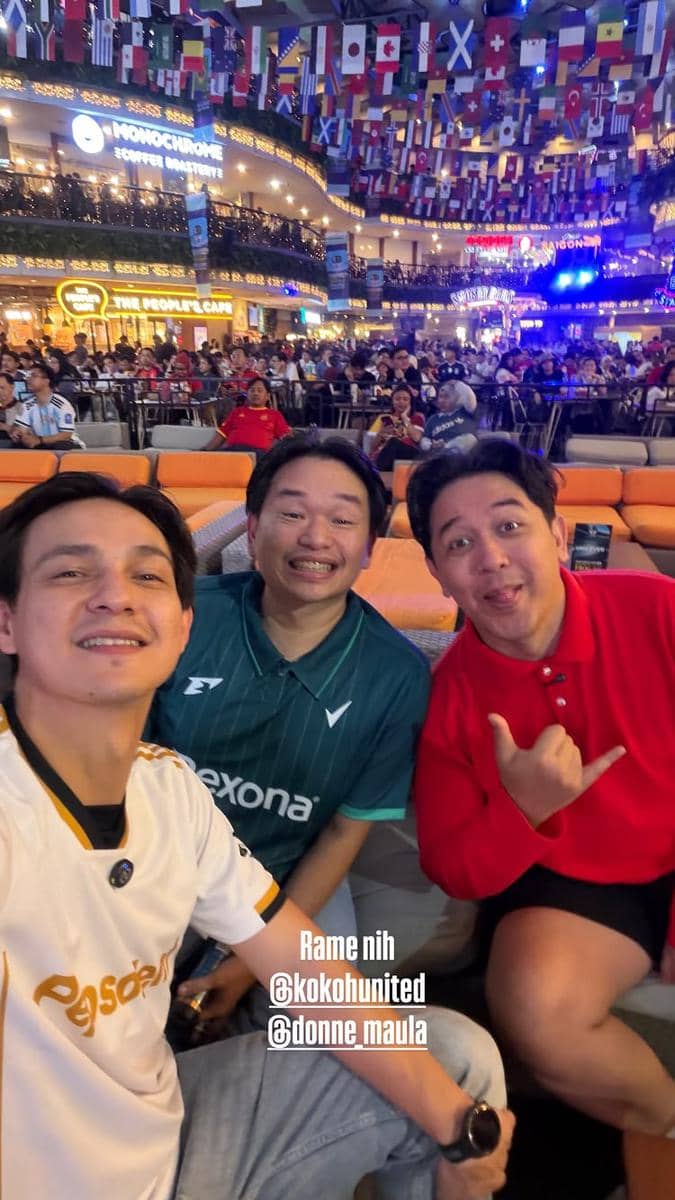 Tiga pria berpose bersama di area nonton bareng final Piala Dunia 2026 dengan suasana ramai dan bendera berbagai negara di atas mereka.