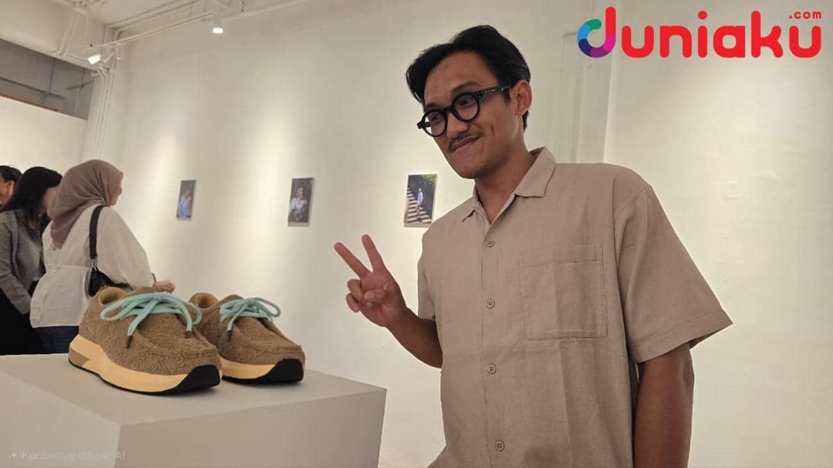 Seorang pria berpose dengan tanda damai di samping sepatu Velocity Moc edisi eye_C yang dipajang di galeri seni.