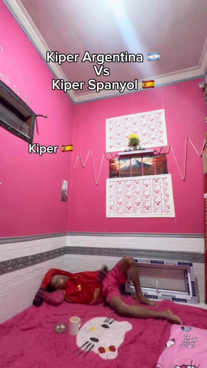 Seorang pria berbaring santai di kamar bernuansa merah muda dengan tulisan meme tentang kiper Argentina dan Spanyol di dinding.