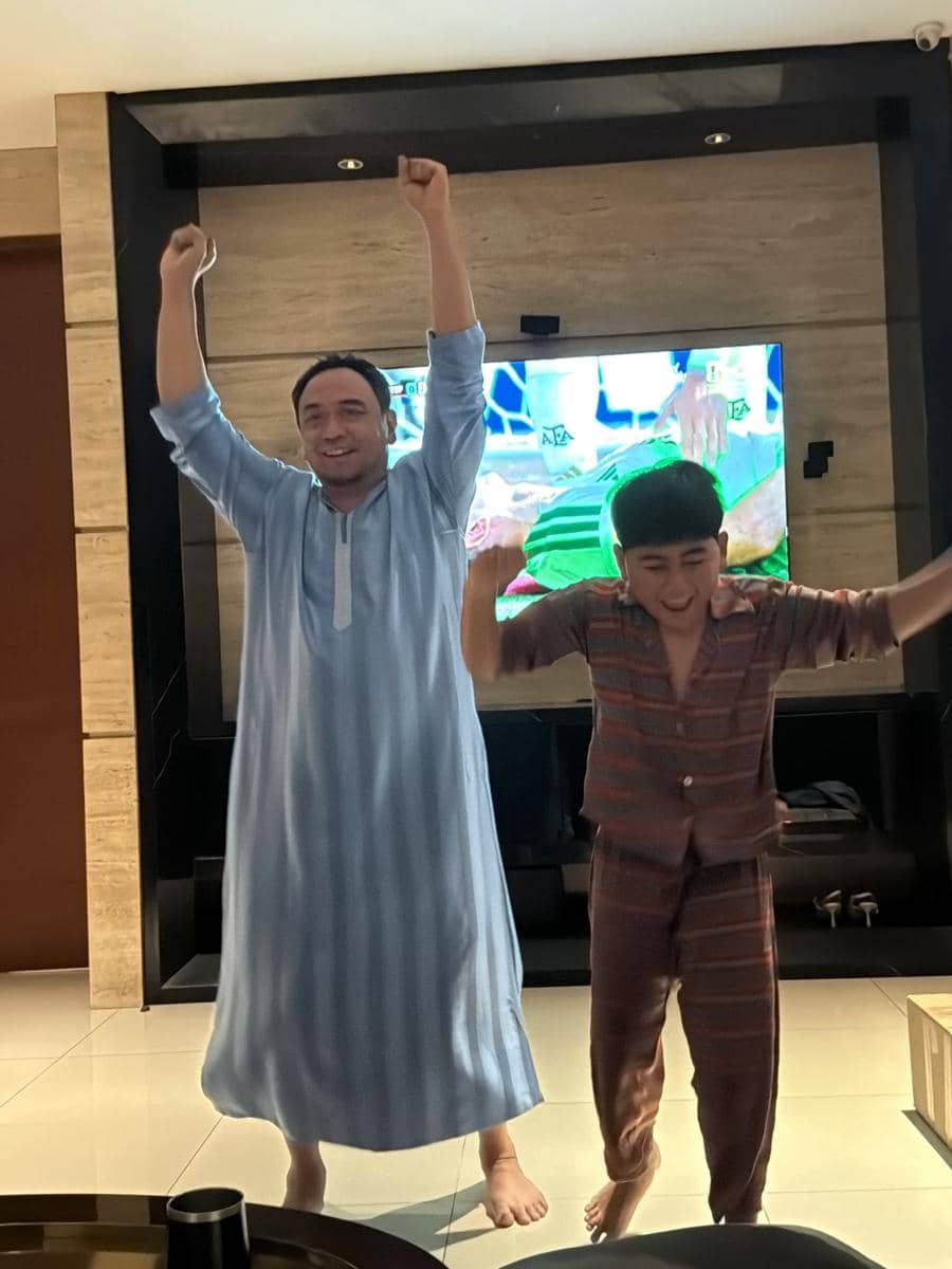 Dua orang bersorak gembira di ruang keluarga sambil menonton pertandingan sepak bola di televisi dengan tangan terangkat ke atas.