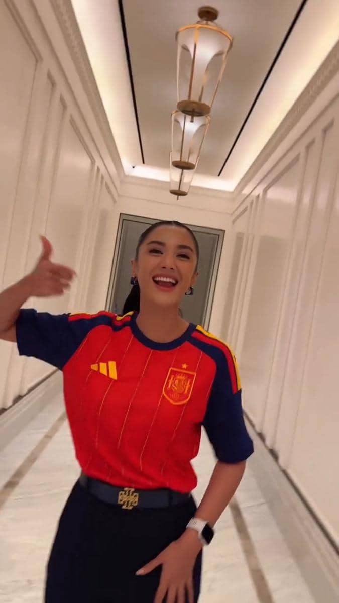 Seorang wanita mengenakan jersey tim nasional Spanyol berwarna merah dan biru tersenyum sambil mengacungkan jempol di lorong bangunan mewah.