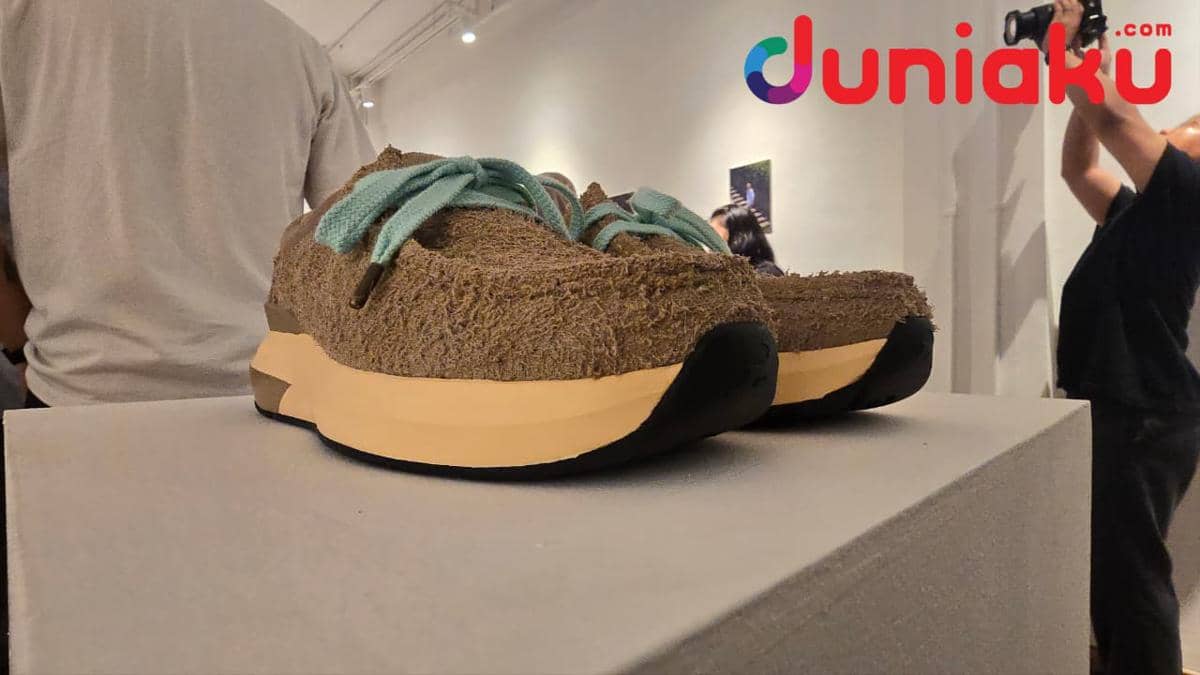 Sepasang sepatu Velocity Moc edisi kolaborasi COMPASS x eye_C dengan bahan suede cokelat dan tali hijau toska dipajang di atas podium.