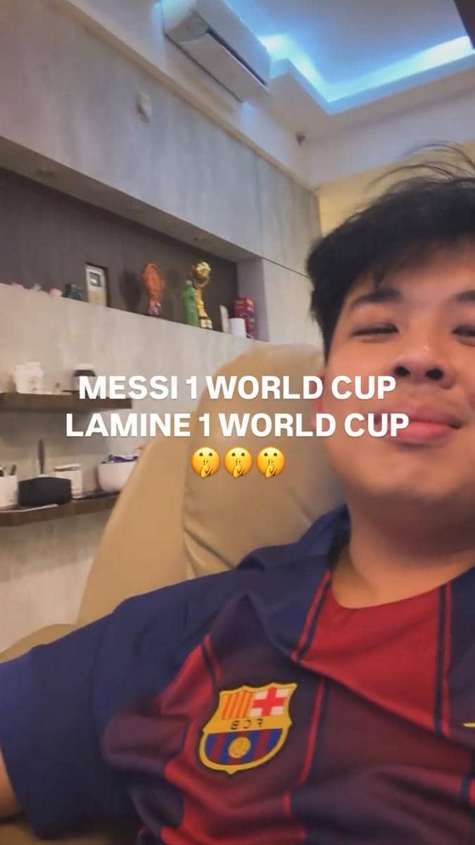 Seorang pria mengenakan jersey Barcelona duduk santai di kursi dengan tulisan di layar membandingkan pencapaian Piala Dunia Messi dan Lamine.