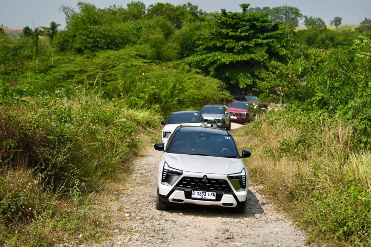 Beberapa unit Mitsubishi New Xforce HEV melintasi jalur bebatuan di area hijau dengan pepohonan dan semak di sekitarnya.