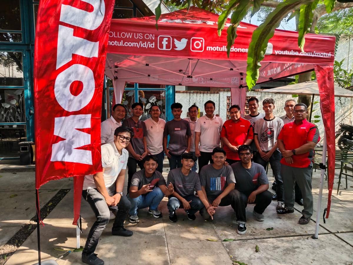 Sekelompok orang berfoto bersama di bawah tenda merah bertuliskan Motul dalam acara kerja sama edukasi dengan bengkel lokal.