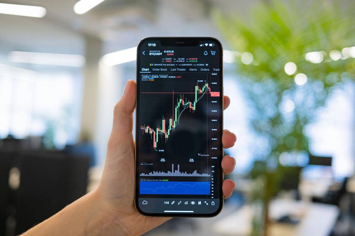 Tangan memegang ponsel yang menampilkan grafik pergerakan harga saham di aplikasi perdagangan dengan latar kantor modern.
