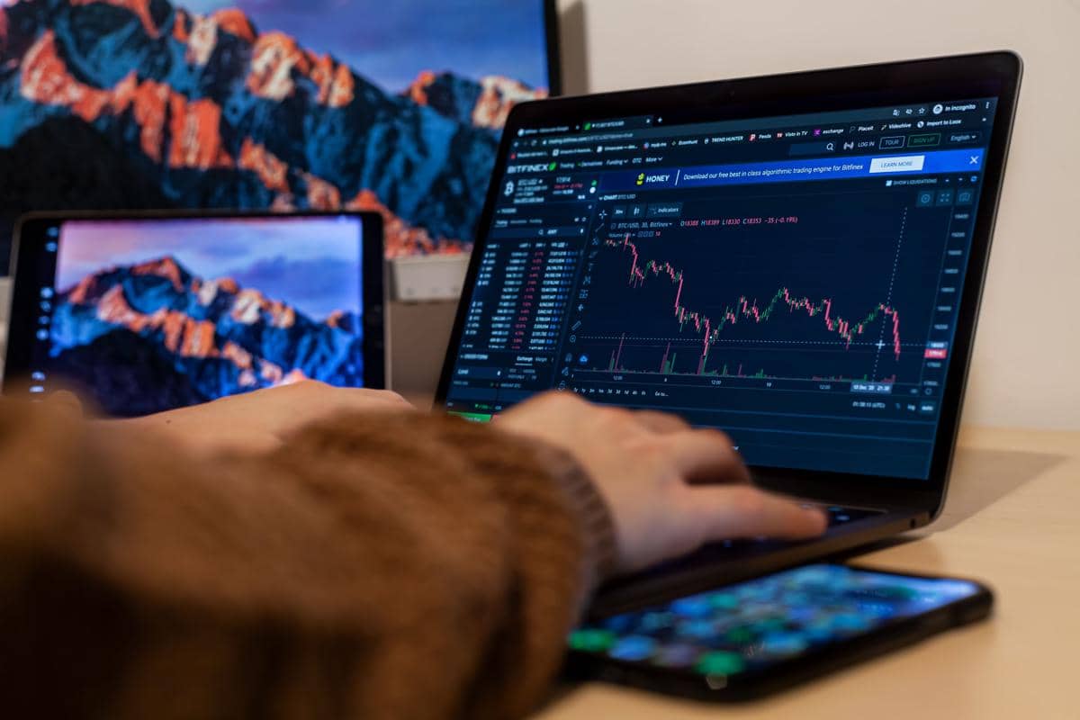 Seseorang menggunakan laptop untuk memantau grafik pergerakan saham di layar dengan latar belakang pegunungan di monitor lain.