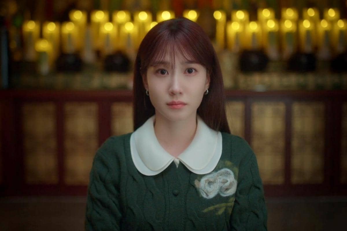 Seorang wanita berambut panjang mengenakan sweter hijau dengan kerah putih duduk di depan latar belakang lilin menyala dalam adegan drama Spooky in Love.
