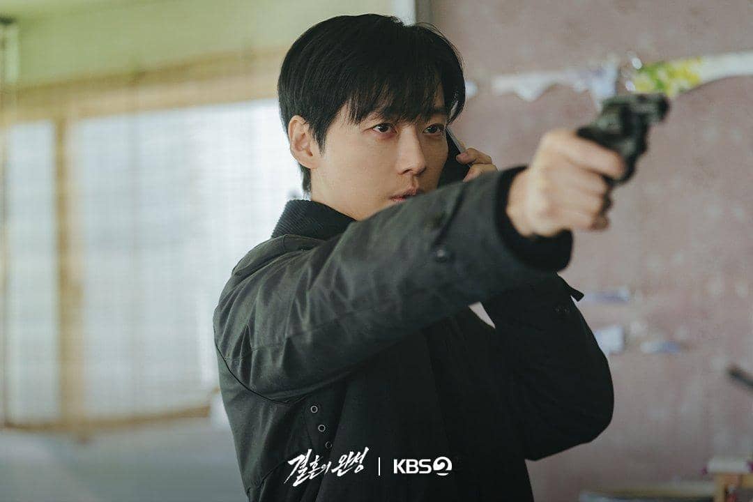 Seorang pria mengenakan jaket gelap sedang mengarahkan pistol sambil memegang ponsel di telinga dalam adegan drama The Husband.