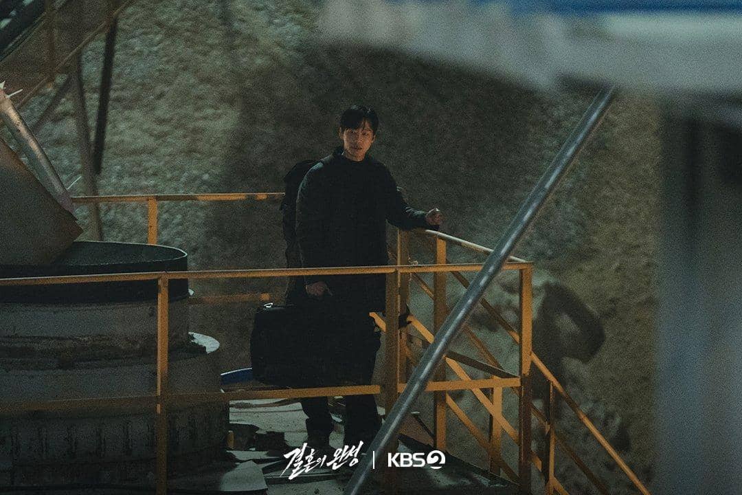 Seorang pria berdiri di tangga logam dengan membawa tas besar pada malam hari dalam adegan drama The Husband produksi KBS2.