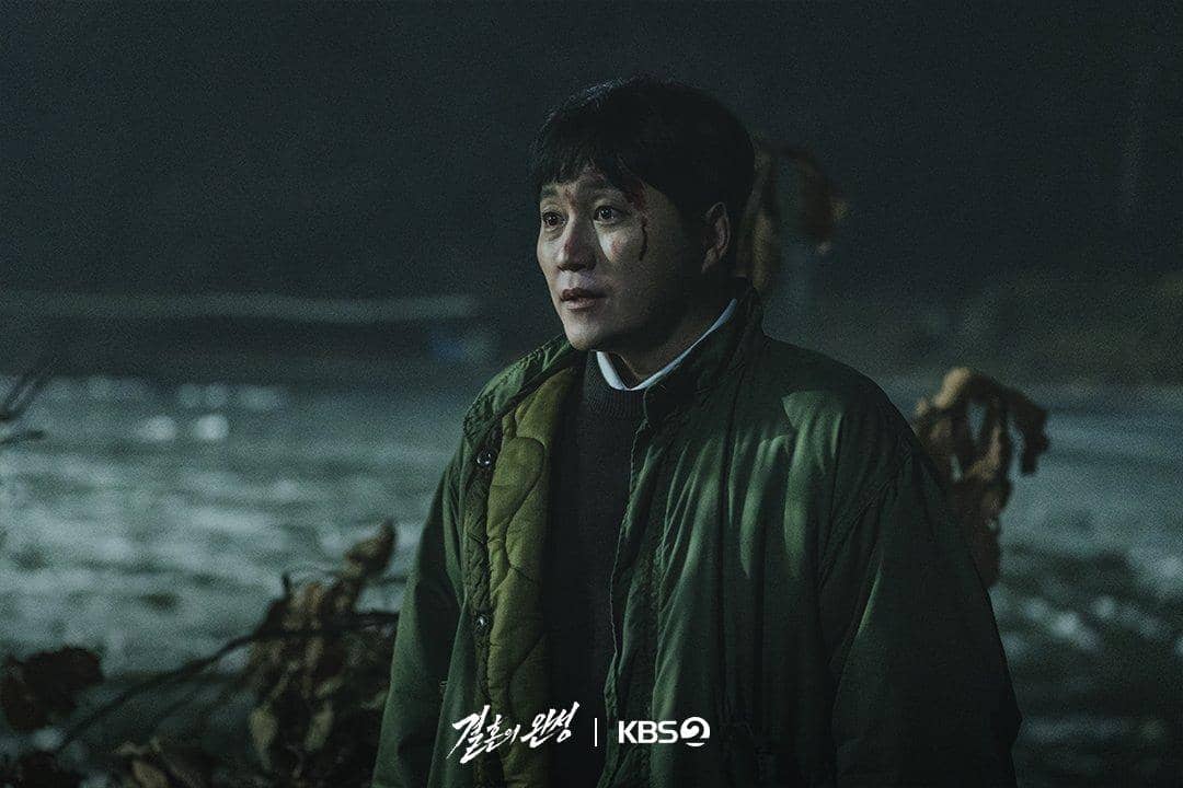 Seorang pria dengan jaket hijau berdiri di luar ruangan pada malam hari dengan luka di wajahnya dalam adegan drama The Husband.
