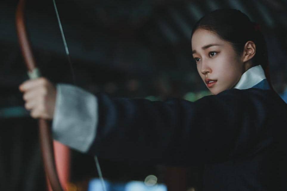 Seorang perempuan mengenakan hanbok gelap sedang membidik dengan busur panah dalam adegan drama The East Palace.