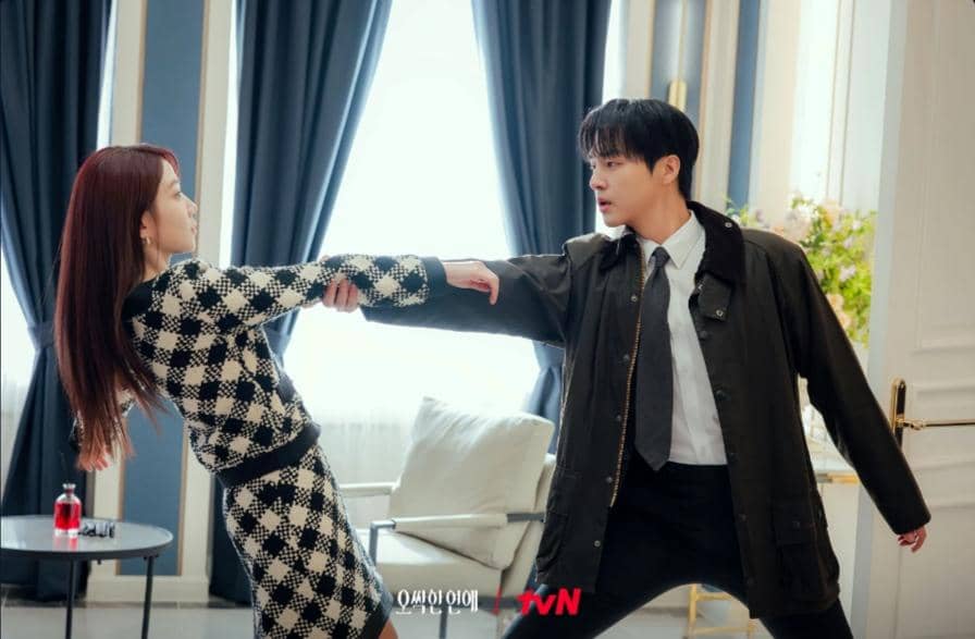 Dua karakter dalam drama Spooky in Love sedang berhadapan di ruang tamu, saling menarik tangan dengan ekspresi tegang.