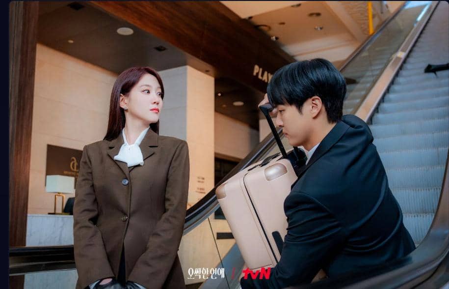 Dua karakter dalam drama Spooky in Love berdiri di eskalator, mengenakan setelan formal dengan suasana hotel atau gedung modern.