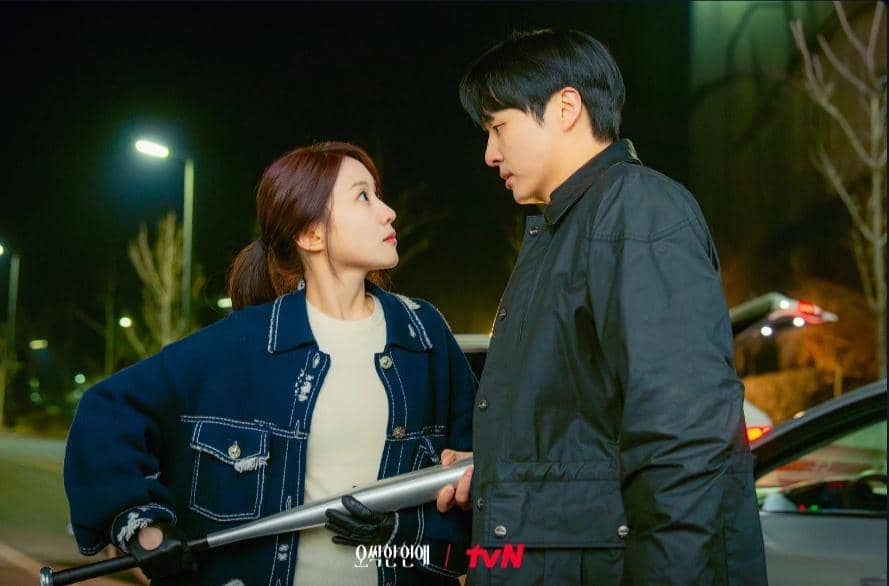 Dua karakter dalam drama Spooky in Love saling menatap intens di malam hari di dekat mobil dengan suasana tegang.